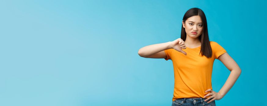 Moody unsatisfied cute asian girl picky, disliking bad restaurant food smirking displeased, grimacing disappointed show thumb down, shaking head negative judgement, stand blue background.