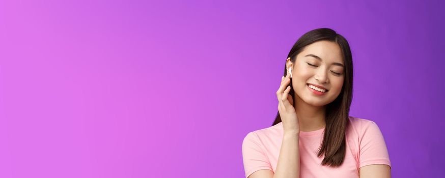 Cute asian girl enjoying listening music, put on wireless headphone, tilt head close eyes dreamy smiling satisfaction, perfect sound, make volume earphone louder, stand purple background.