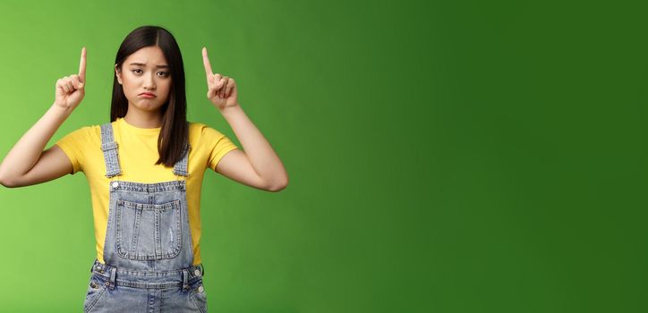 Pity cute sad asian girl brunette, grimacing pulling sorrow upset face frowning bothered, pointing fingers up regret awful situation, indicate top promo, stand green background unhappy.