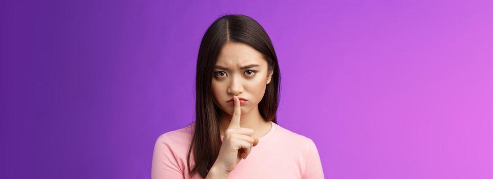 Close-up offended serious-looking upset asian young girl frowning cannot focus disturbed loud conversation, hush stare camera unsatisfied, show shush sign press finger lips, purple background.
