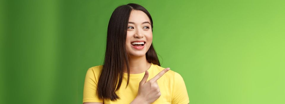 Close-up cheerful friendly asian outgoing girl having fun, pointing turn left amused, lively talking, discuss friends interesting event, stand green background chatting carefree entertained.