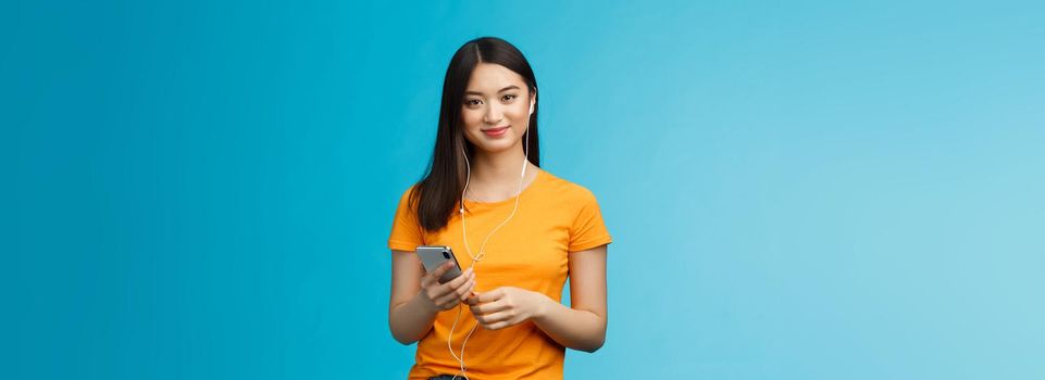 Carefree attractive pleasant, tender asian urban woman wear earphones, listen music via headphones hold smartphone, smiling delighted, found awesome new song, stand blue background.