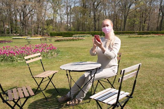 young woman in a beige jumper and a protective mask takes a selfie in the park against the backdrop of blooming tulips in early spring social media using a smartphone, High quality photo