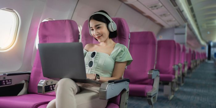 Joyful asian woman sits in the airplane using laptop and listen music while go to travel.