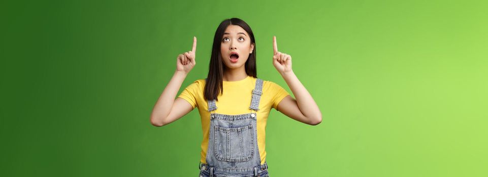 Omg look shocking price fall. Stunned speechless amazed asian girl drop jaw, wondering what happening upstairs, pointing lookin up impressed, staring surprised stand green background.