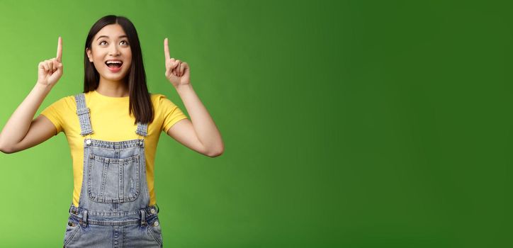 Carefree amused asian brunette in yellow t-shirt dungarees, smiling amazed and fascinated, look pointing up, observe cool new promo, grinning excited, enjoy watching top copy space.