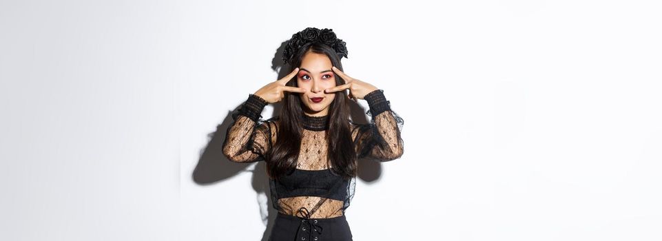 Beautiful asian woman in halloween party outfit looking at upper left corner, making peace gestures over eyes, standing in gothic lace dress with black wreath over white background.
