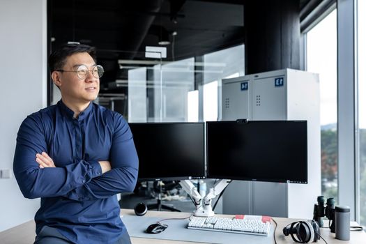 Portrait of Asian businessman, man in glasses and shirt looking out window and smiling, worker inside office building with crossed arms.