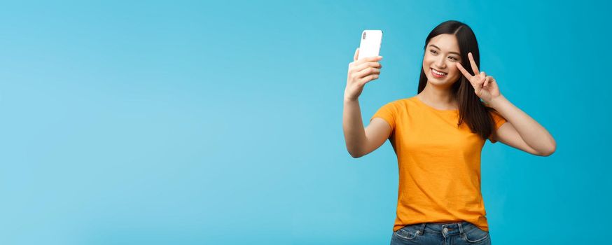 Friendly cheerful pretty asian woman brunette short haircut taking selfie, smiling joyfully hold smartphone show peace, victory sign at phone screen front camera, talking family video-call.