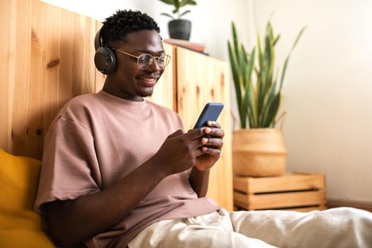 Young african american man with glasses relaxing at home listening to music with headphones while using mobile phone. Copy space. Lifestyle.