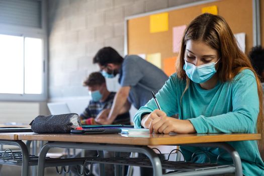 High school female student wearing face mask in class.doing homework. Teacher helping male teen student in background. Education concept. Healthcare concept.