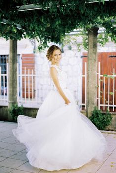 Bride stands on the tiled floor of a pergola, holding up the skirts of her puffy dress. High quality photo