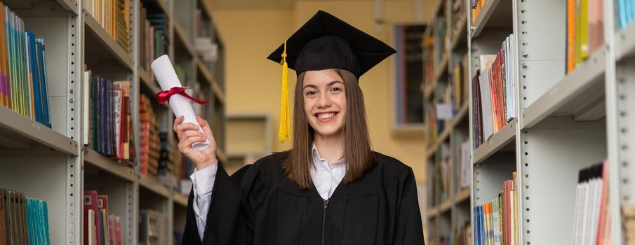 Happy young woman in graduate gown holding diploma in the library. Widescreen