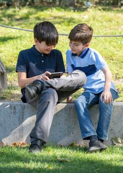 Cute two boys sitting on the park and play online games. One boy holding the smartphone. Young boys use the phone.