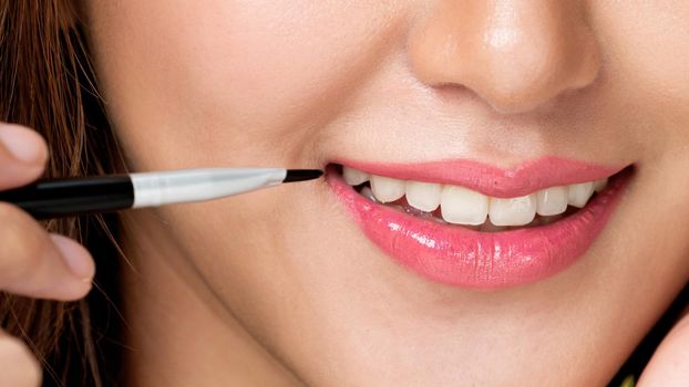 Close up young ardent woman with healthy fair skin applying lipstick on her lip while looking at camera. Beauty concept with cosmetic and makeup.