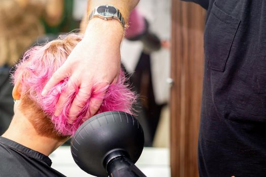 Drying short pink bob hairstyle of a young caucasian woman with a black hair dryer with the brush by hands of a male hairdresser in a hair salon, close up