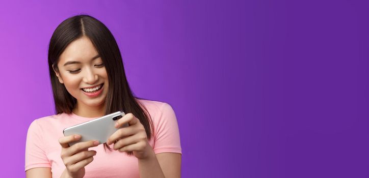 Close-up joyful attractive asian woman brunette having fun spend time playing smartphone game, laughing smiling eager win race, hold phone horizontal beating score, purple background.