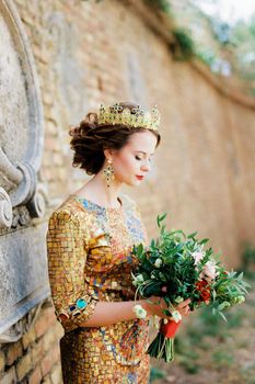 Bride in a crown with a bouquet of flowers stands near a stone wall. High quality photo