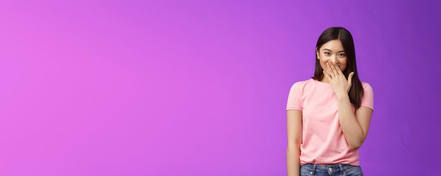 Timid cute stylish modern asian woman giggling, gossiping, cover smiling lips, laughing out loud joyfully, look camera entertained, fool around, spread funny rumors, stand purple background.