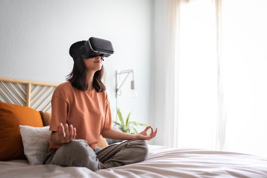 Young asian woman meditating with help of VR experience app sitting on bed. Asian girl doing meditation using virtual reality goggles at home. Copy space. Technology and lifestyle concepts.
