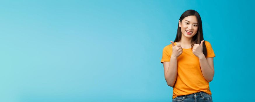 Supportive cute friendly asian girl give thumbs up and smiling, cheering for you, congratulate nice work, approving good idea, like nice outfit, grinning glad accept plan, stand blue background.