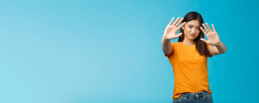 Asian moody displeased silly timid girl asking turn off light not photograph her, raise hands block, cover face step back and cringe disappointed, standing blue background upset reluctant. Copy space