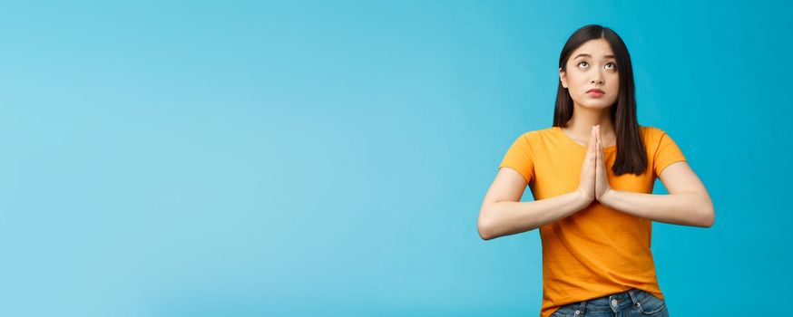 Serious-looking hopeful cute asian woman begging god hear prayers, supplicating, making wish look up sky talking lors, believe have faith, stand blue background pleading good fortune luck.