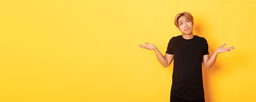Portrait of clueless asian blond guy, wearing black clothes, shrugging and looking puzzled, standing yellow background.