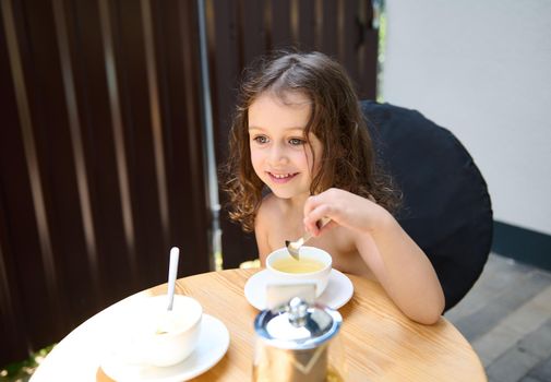 Adorable Caucasian little girl sitting at a table in the urban summer terrace, drinking healthy antioxidant refreshing and delicious herbal and fruit tea and smiling a cheerful toothy smile