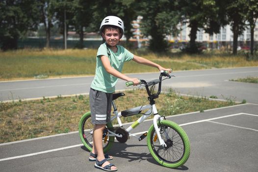 Charming active Caucasian child boy, in protective helmet, looking at camera, standing near his bicycle on the asphalt road outdoor. Physical activity. Active lifestyle. Equilibre and balance. Sport