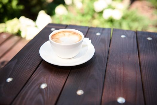 Still life with a white cup of frothy cappuccino with foam on a wooden table, in the summer terrace of a cafeteria. Food and drink