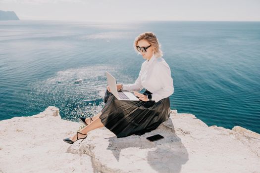 Successful business woman in yellow hat working on laptop by the sea. Pretty lady typing on computer at summer day outdoors. Freelance, travel and holidays concept.