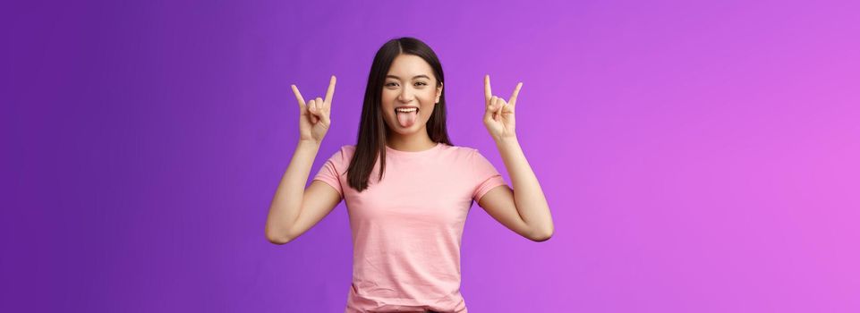 Rebellious amused asian girl enjoy rock-n-roll, attend awesome concert have fun, show tongue make heavy-metal gesture, express excitement and joy, stand purple background cheerful.