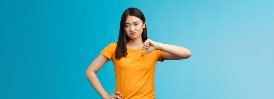 Moody unsatisfied cute asian girl picky, disliking bad restaurant food smirking displeased, grimacing disappointed show thumb down, shaking head negative judgement, stand blue background.