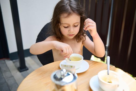 Beautiful Caucasian little girl, sitting at a table and using a tea spoon, stirring sugar in the white teacup with healthy antioxidant herbal tea