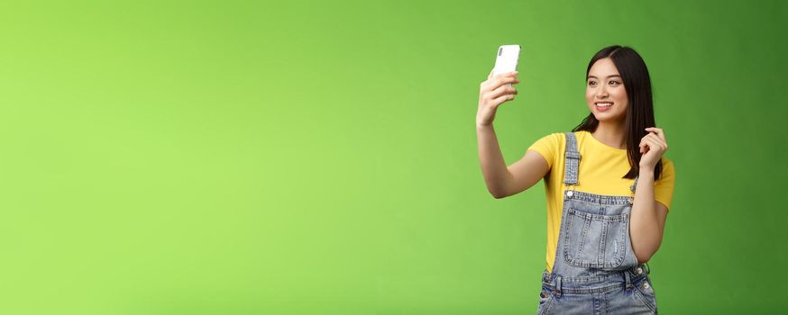 Glamour tender teenage asian female taking selfie, checking haircut, look smartphone front camera, photographing, record video message post online, smiling lovely, green background.