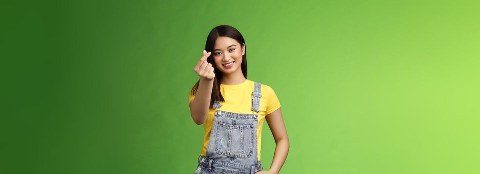 Lovely tender carefree asian girlfriend show korean love sign, make finger heart smiling cute, showing passion and affection, stand green background in overalls and yellow t-shirt, express sympathy.