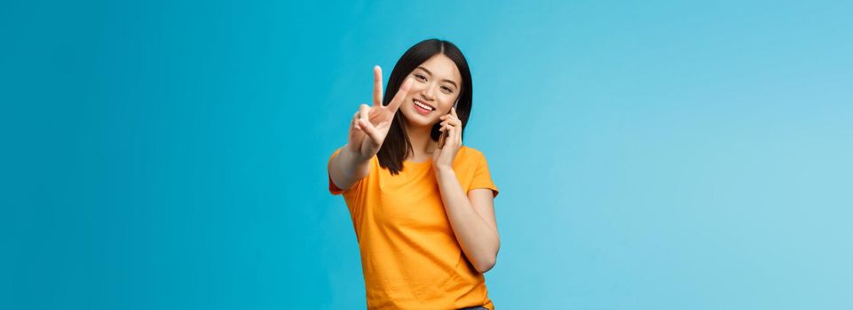 Charismatic cute girl share positive news talking smartphone show peace, victory sign smiling satisfied, standing blue background receive pleasant phone call, achieve deal via phone-call.