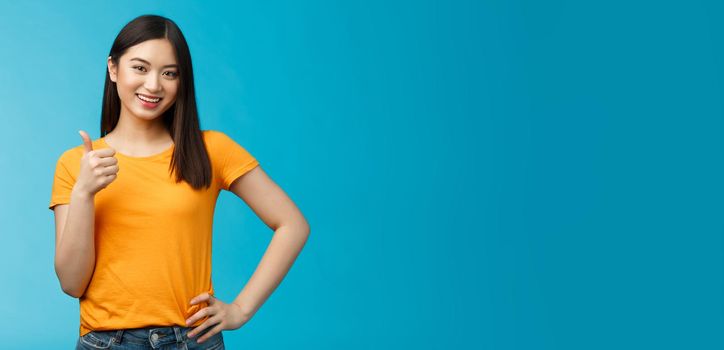 Confident cheerful asian brunette girl show thumb-up satisfied, give approval, liking good choice, smiling delighted, accept your plan, positive judgement, stand blue background. Copy space