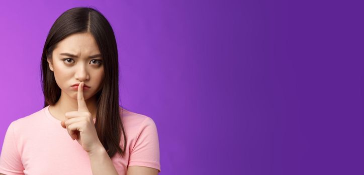 Close-up offended serious-looking upset asian young girl frowning cannot focus disturbed loud conversation, hush stare camera unsatisfied, show shush sign press finger lips, purple background.