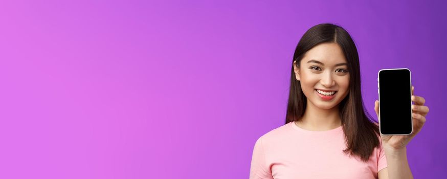 Close-up attractive pleasant female shop assistant present new phone app, hold smartphone show telephone screen, introduce application on display, stand purple background delighted.