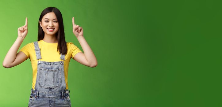 Friendly cute good-looking asian brunette female pointing up, raise fingers top advertisement, self-assured showing promo, smiling satisfied, introduce new product, green background.