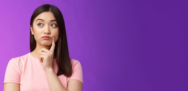 Close-up thoughtful cute asian girl dreamy standing purple background, touch cheek, turn left, thinking, doubting how act, wondering information, contemplating view from window.