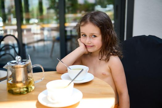 Caucasian cute little girl sitting at a table in the urban summer terrace, drinking healthy antioxidant refreshing fruit tea