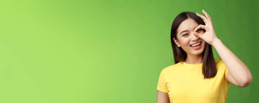 Close-up friendly carefree outgoing nice asian girl brunette having fun, tilt head joyfully, show okay ok sign look through circle amused, amazed perfect quality product, stand green background.
