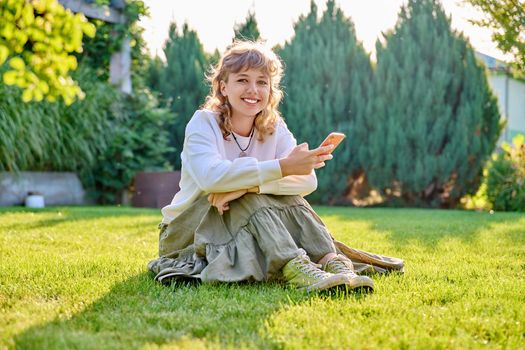 Portrait of smiling teenage blonde curly female with smartphone in hands, looking at camera sitting on grass in backyard, lawn in park. Adolescence, technology, beauty, lifestyle, youth concept