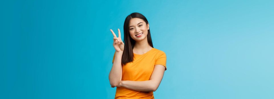 Optimistic cute tender asian young millennial girl send positive vibes, tilt head show peace, victory sign, smiling broadly, wish fantastic holidays, stand blue background joyful upbeat.