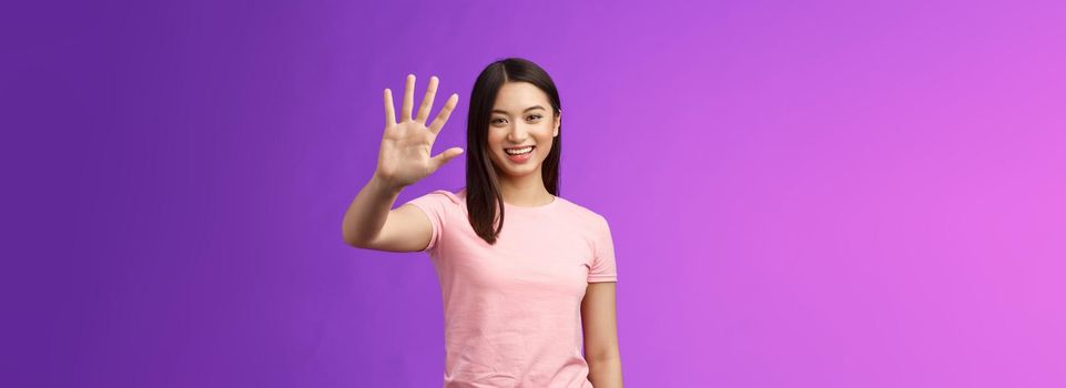 Friendly cute asian tender girl raise palm waving goodbye, smiling joyfully, farewell, gladly welcome friends inviting come in, saying hi hello, greeting, stand purple background. Copy space