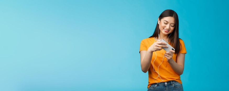 Cute entertained good-looking urban female student spend break playing racing game, hold smartphone horizontal tilt phone move car, smiling broadly standing enthusiastic blue background.