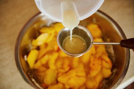 Top view of pouring lemon juice into a metal bowl with sliced peaches while preparing homemade peach confiture. Canning peach. Preparing delicious jam